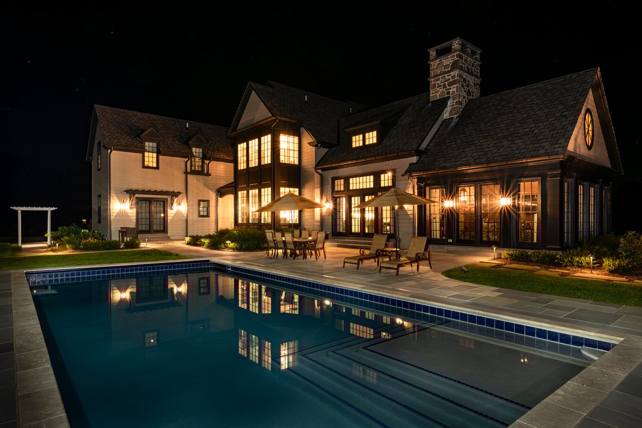 Grand backyard view with glowing pool and seating spaces designed with custom hardscaping, landscaping and lighting in Wilmington, DE