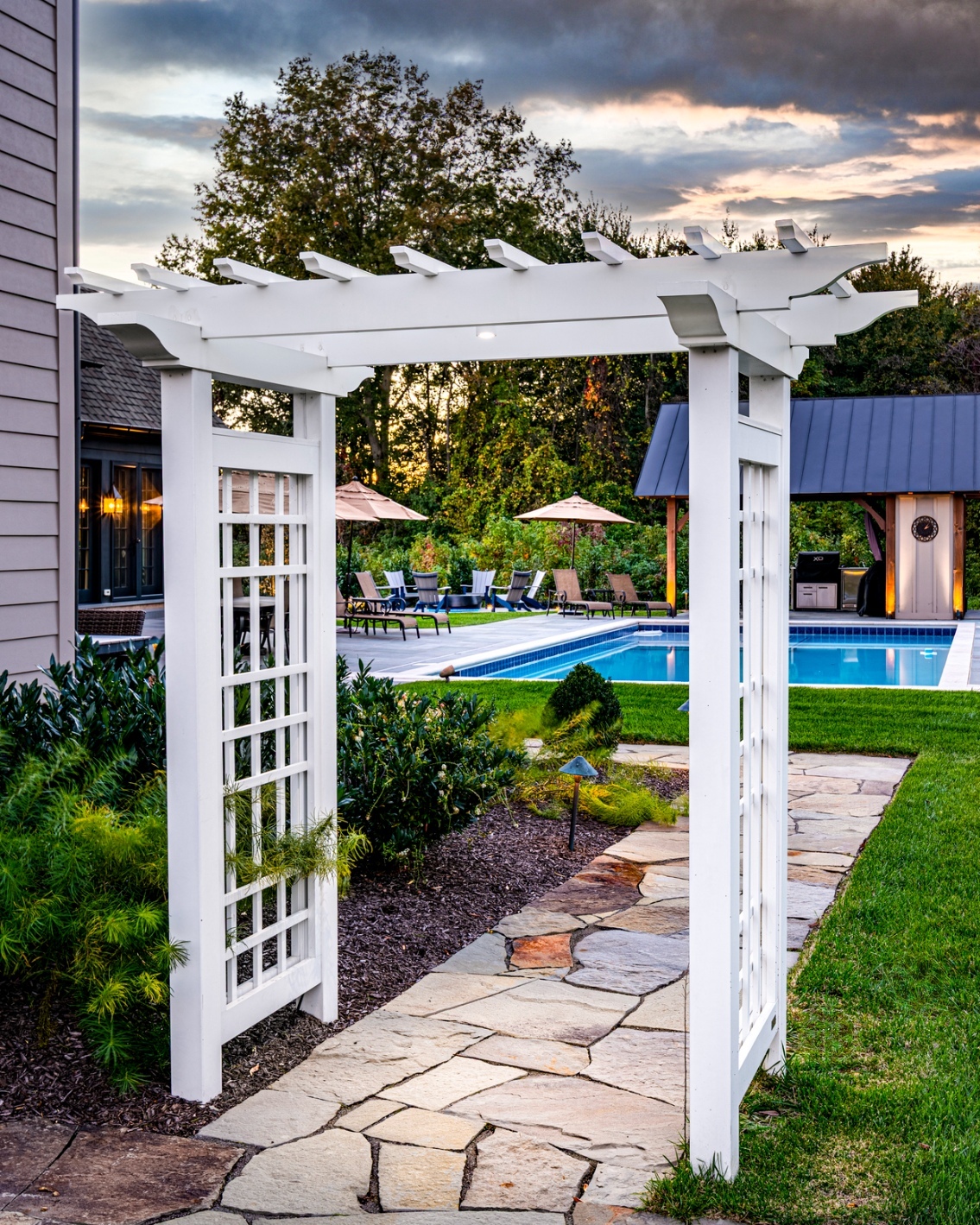 Stone walkway and white arbor pergola framing pool views with custom hardscaping, landscaping and lighting in Wilmington, DE