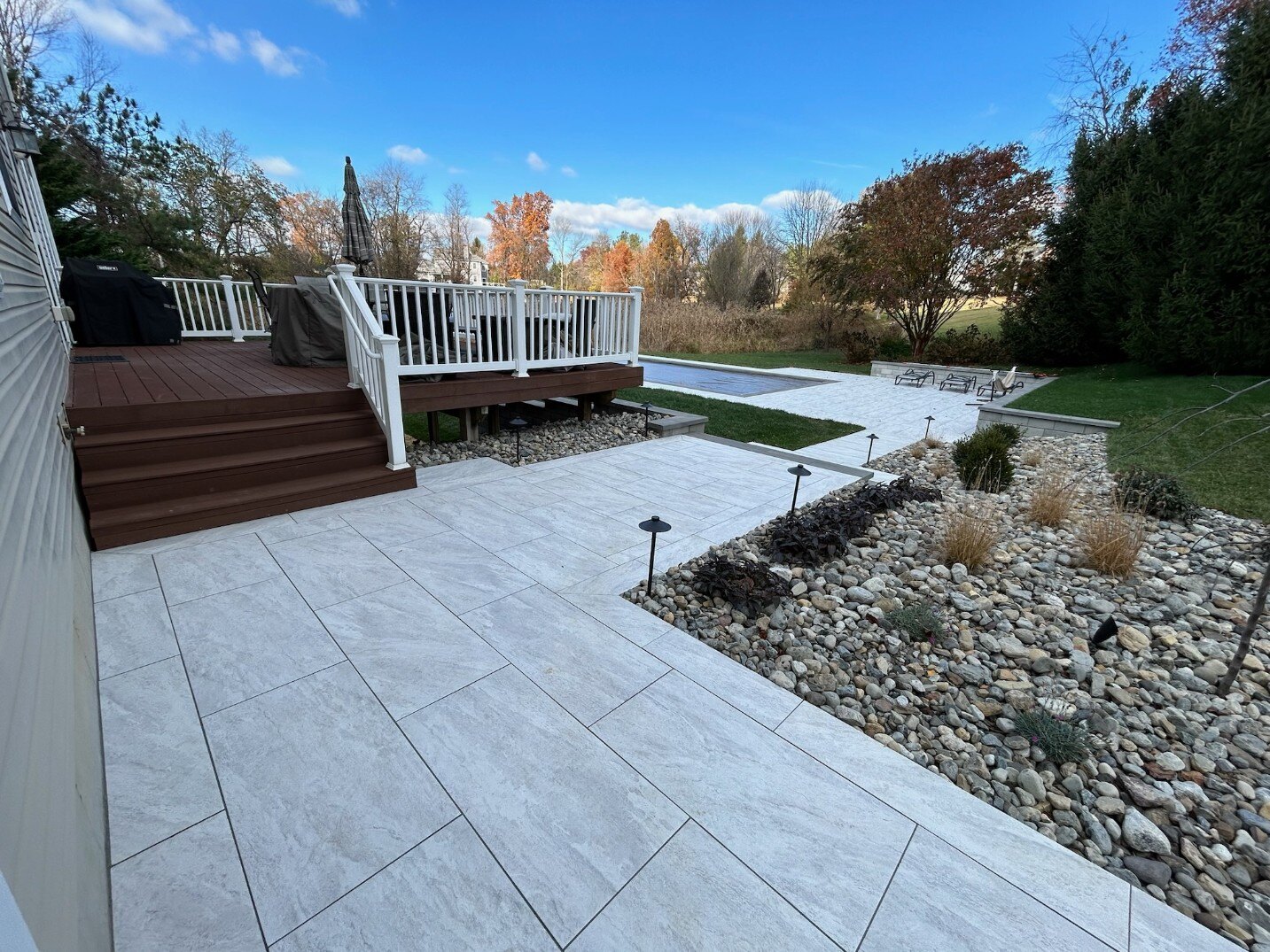 A spacious backyard patio with porcelain pavers, a wooden deck, and a stunning pool in Newark, DE by First Class Lawn Care.
