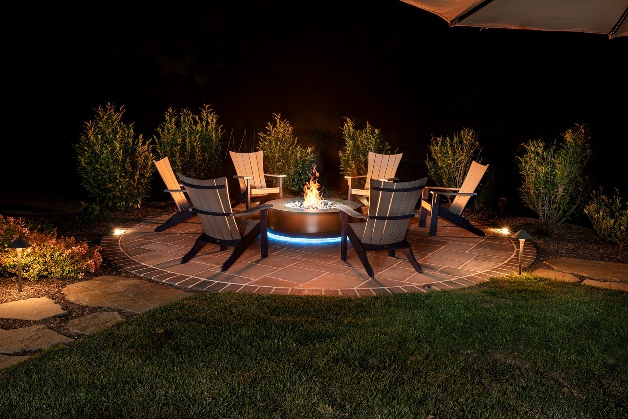 Circular fire pit with Adirondack chairs and surrounding landscape featuring custom hardscaping, landscaping and lighting in Wilmington, DE