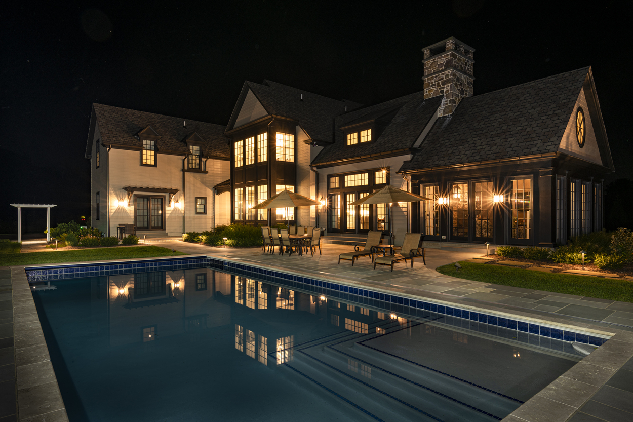 Grand backyard view with glowing pool and seating spaces designed with custom hardscaping, landscaping and lighting in Wilmington, DE